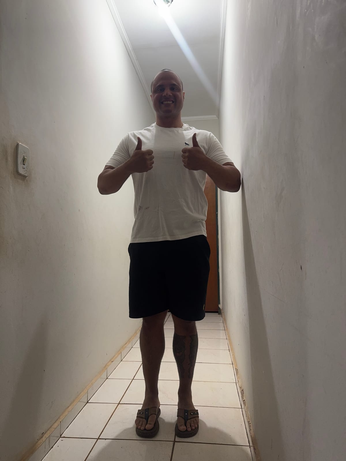 Homem de camiseta branca e bermuda preta, de pé em um corredor estreito de paredes claras e piso de cerâmica, fazendo joia com as duas mãos.